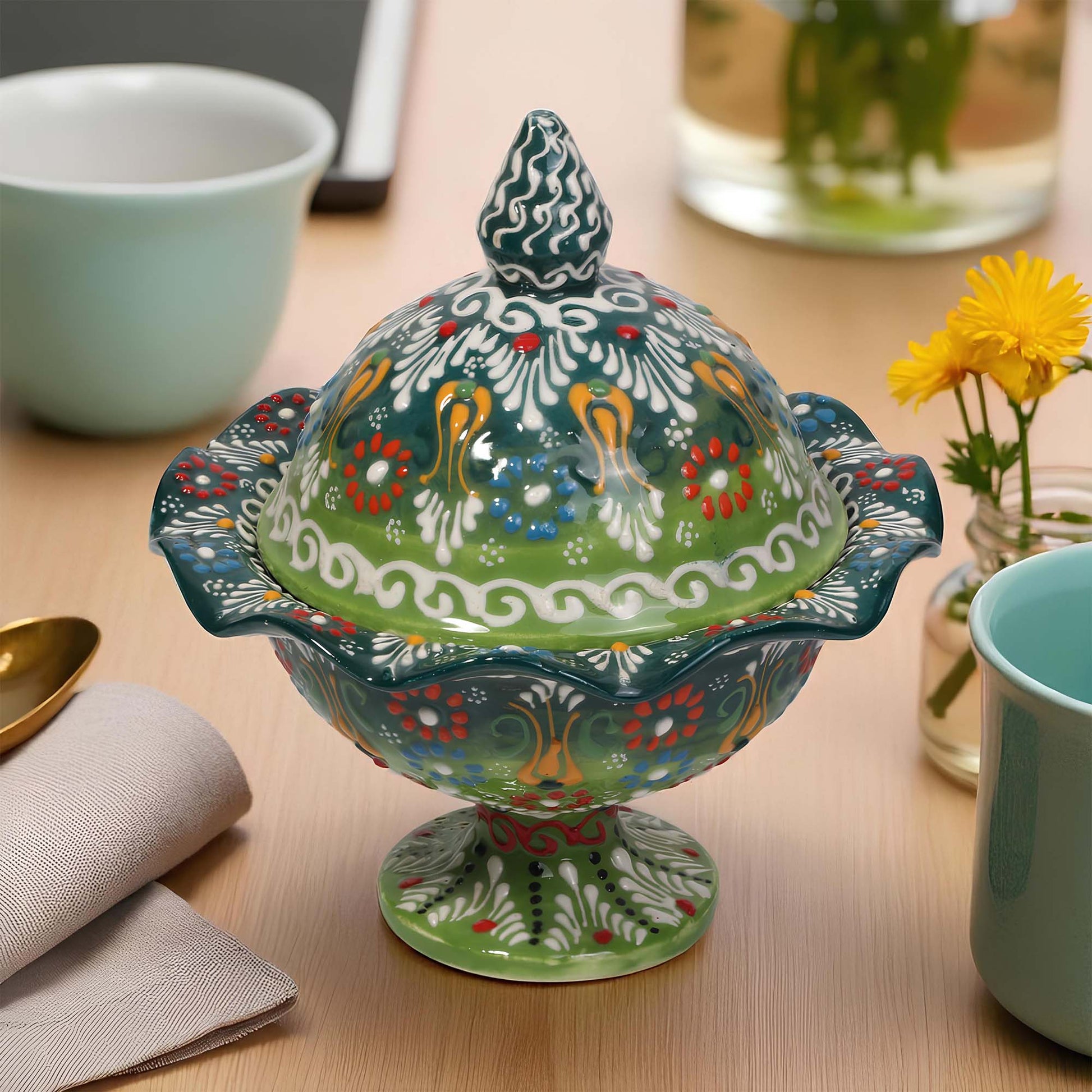 Decorative ceramic sugar bowl with a colorful lid on a table with cups and flowers.