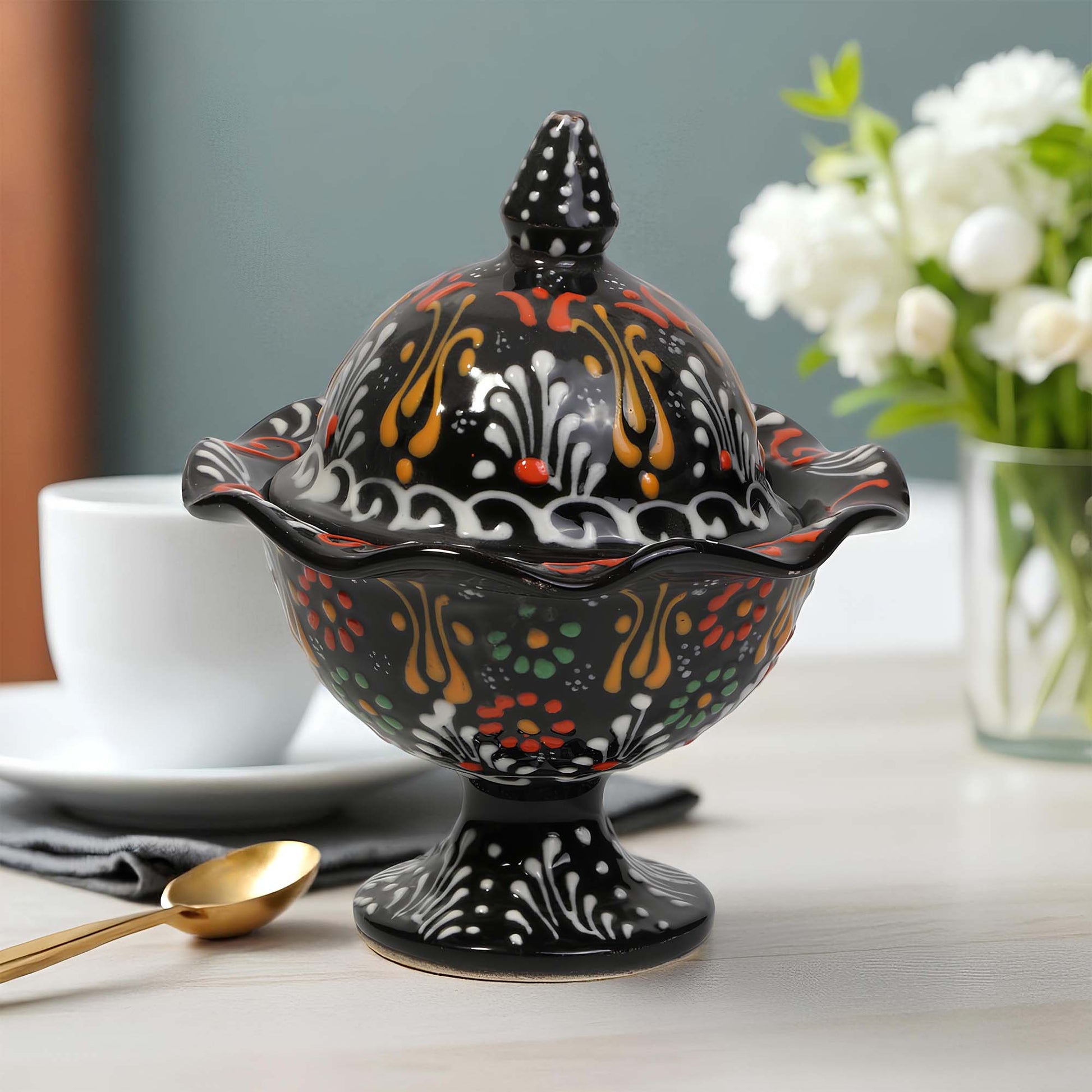 Decorative black ceramic sugar bowl with colorful patterns on a table with a cup and flowers in the background.