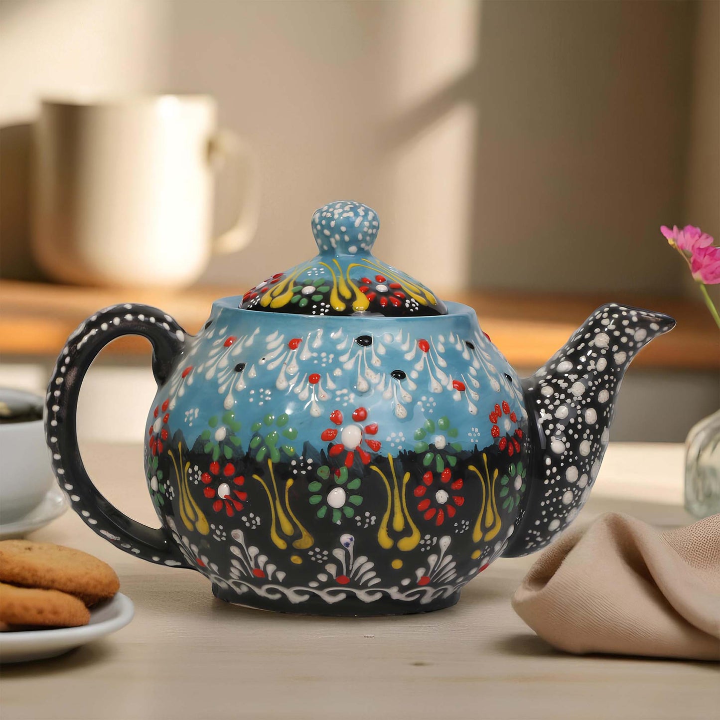 Decorative teapot with floral patterns on a table with cookies and a cup.