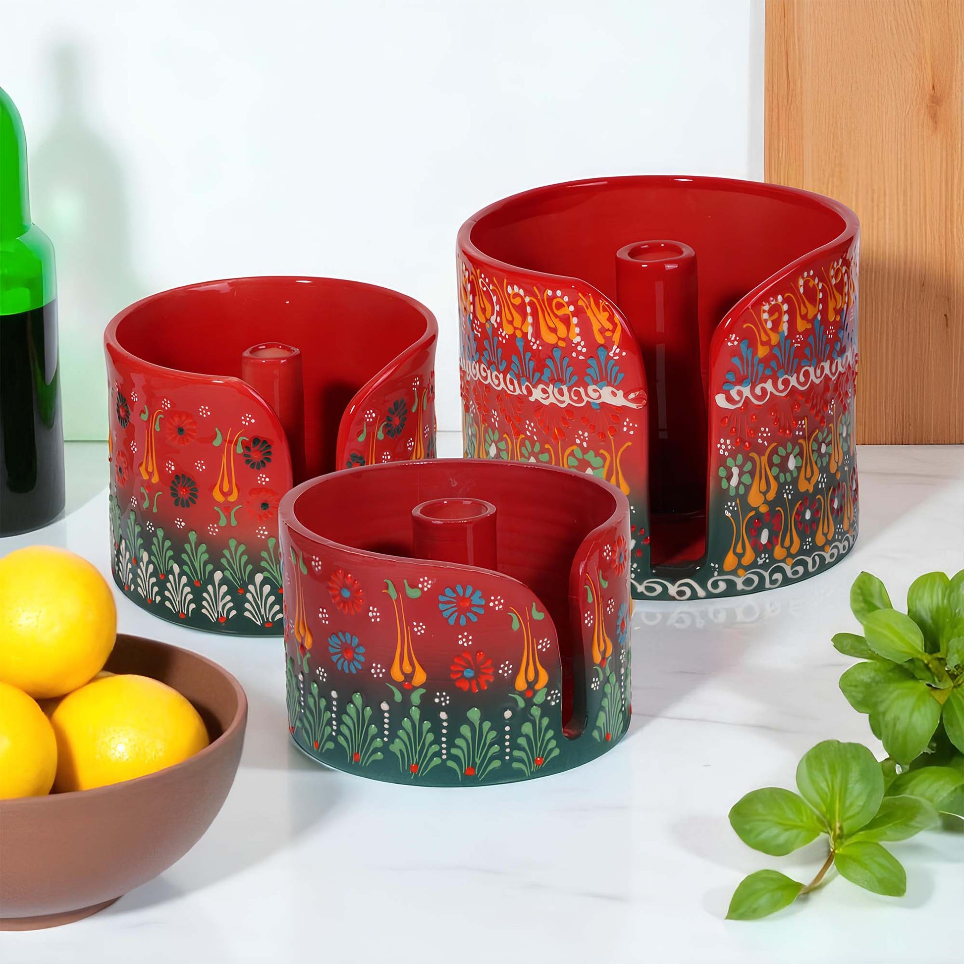 Set of red ceramic paper towel holders with colorful patterns on a white surface with a bowl of lemons and a plant.
