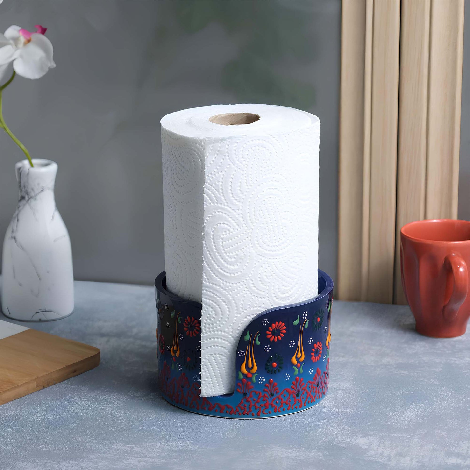 Roll of paper towels in a decorative paper towel holder on a kitchen counter with a vase and cup in the background.