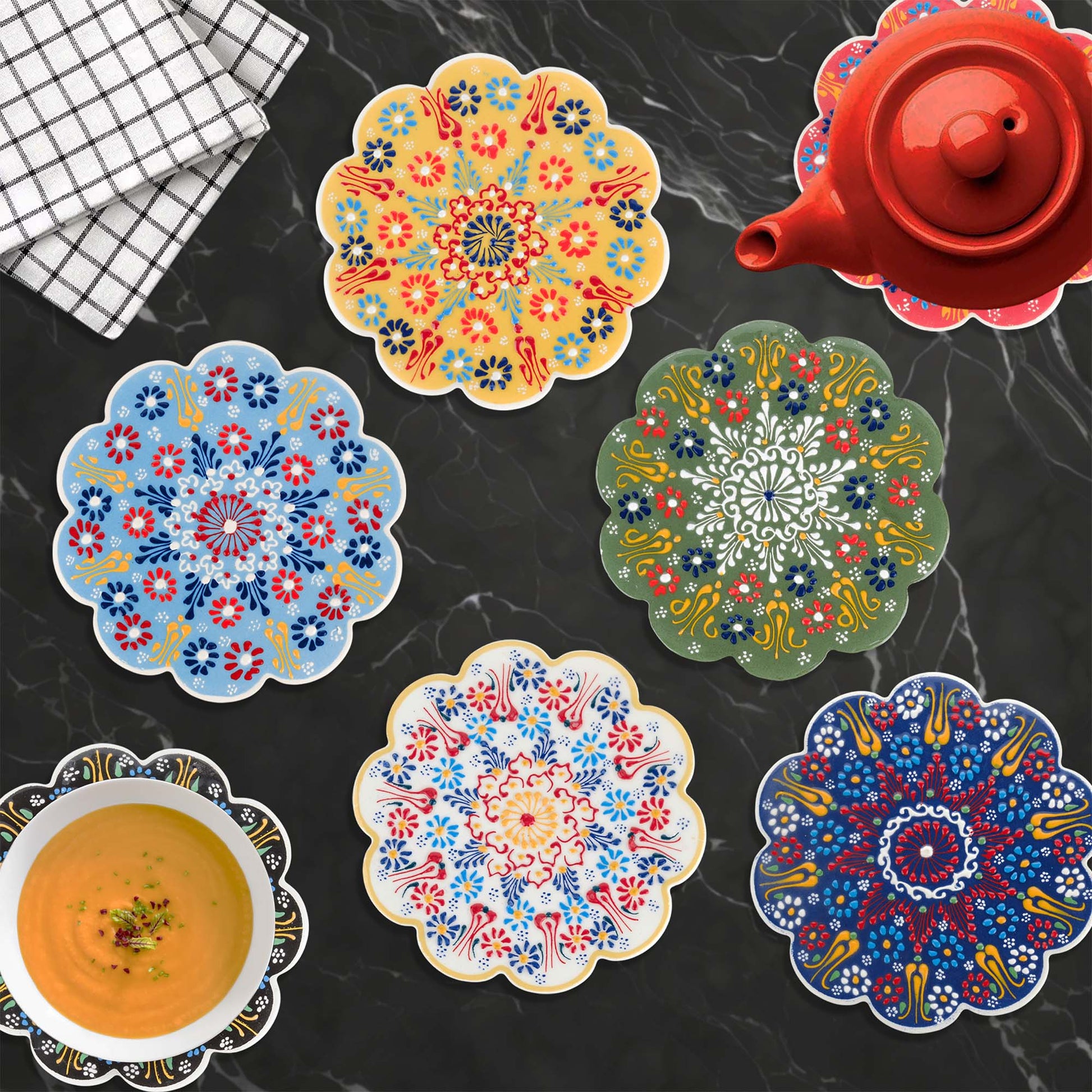 Colorful floral trivets on a dark surface with a red teapot and checkered cloth.