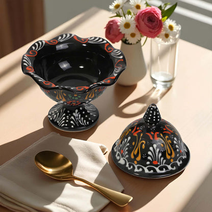 Decorative black sugar bowl with colorful patterns on a table with a gold spoon, white napkin, and flowers.