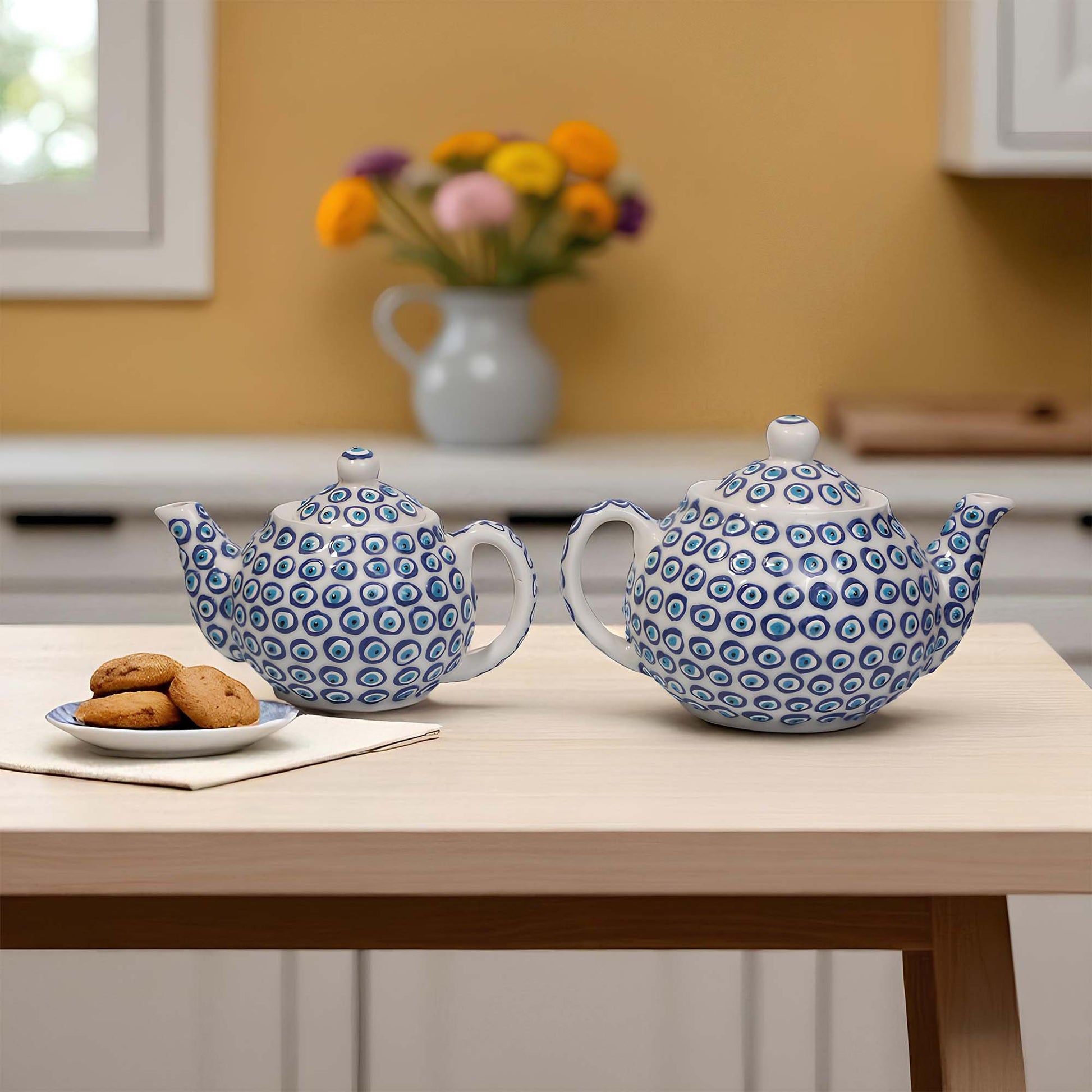 Two blue and white patterned teapots on a wooden table with cookies and a vase in the background.
