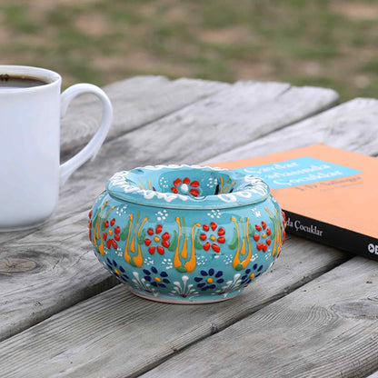 Decorative ceramic ashtray with floral patterns on a wooden surface next to a white mug.