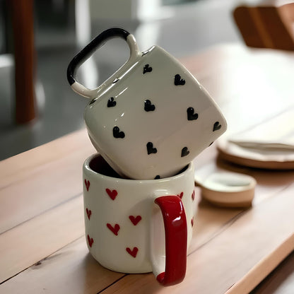 Two ceramic coffee mugs stacked on top of each other, one with a white base and black hearts pattern, and the other with a red handle and white base with red hearts pattern.