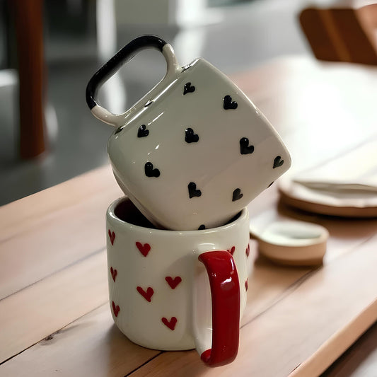 Two ceramic coffee mugs stacked on top of each other, one with a white base and black hearts pattern, and the other with a red handle and white base with red hearts pattern.